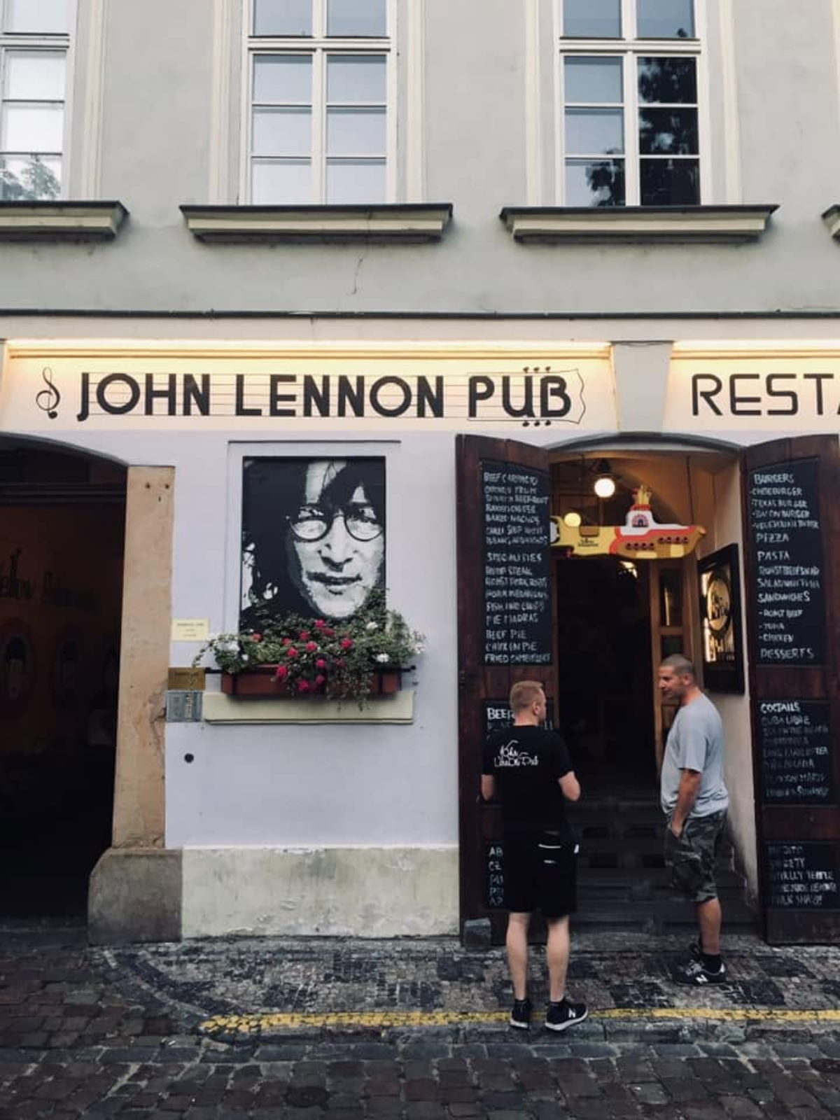 The exterior of the John Lennon Pub in Prague, featuring a mural portrait of John Lennon, hanging flower boxes, and a small yellow submarine detail above the entrance.