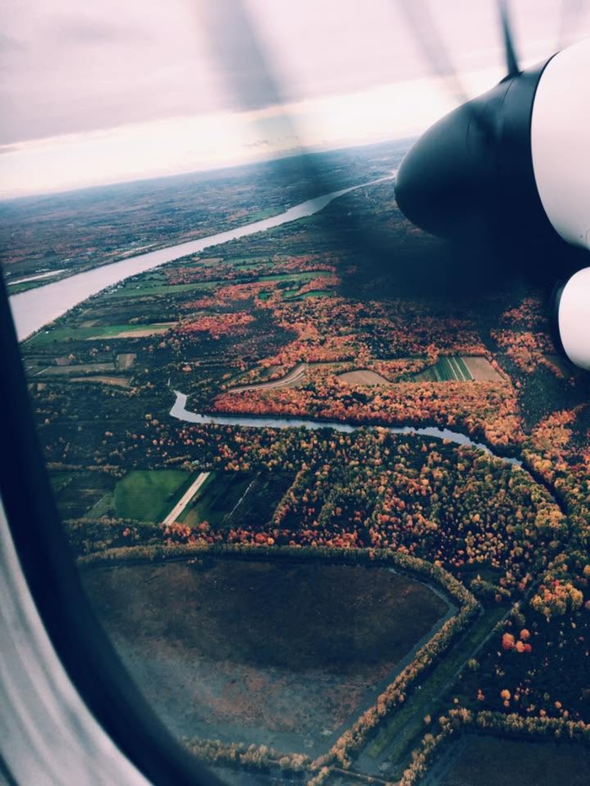 An aerial view from an airplane window over New Brunswick, Canada, showing a winding river cutting through forests in full autumn color.