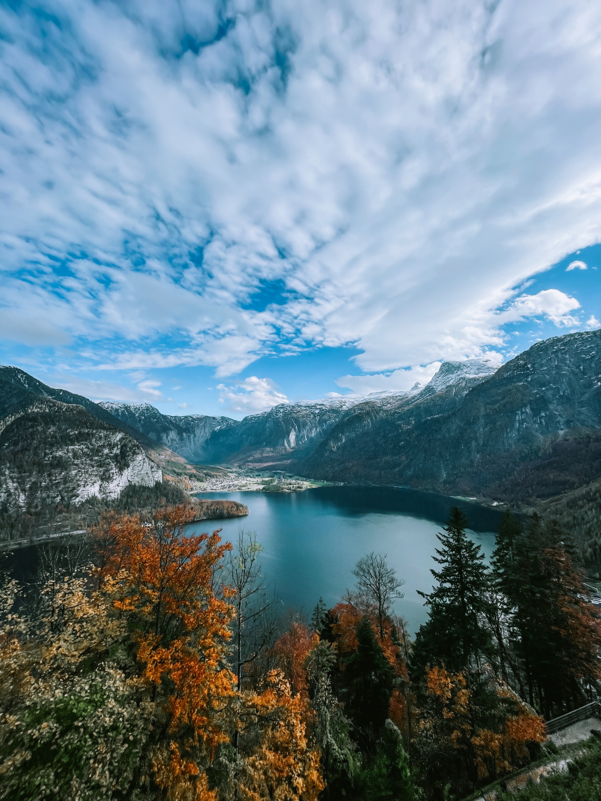 An aerial view of Hallstatt nestled between autumn forests, a deep alpine lake, and snow-dusted mountains.