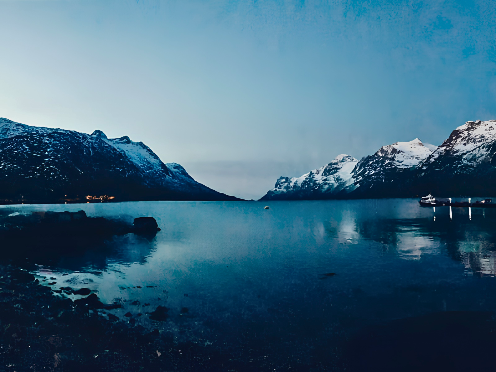 A deep blue twilight over a mirror-still Tromsø fjord, snow-capped mountains rising on either side and a lone boat moored at the shore.