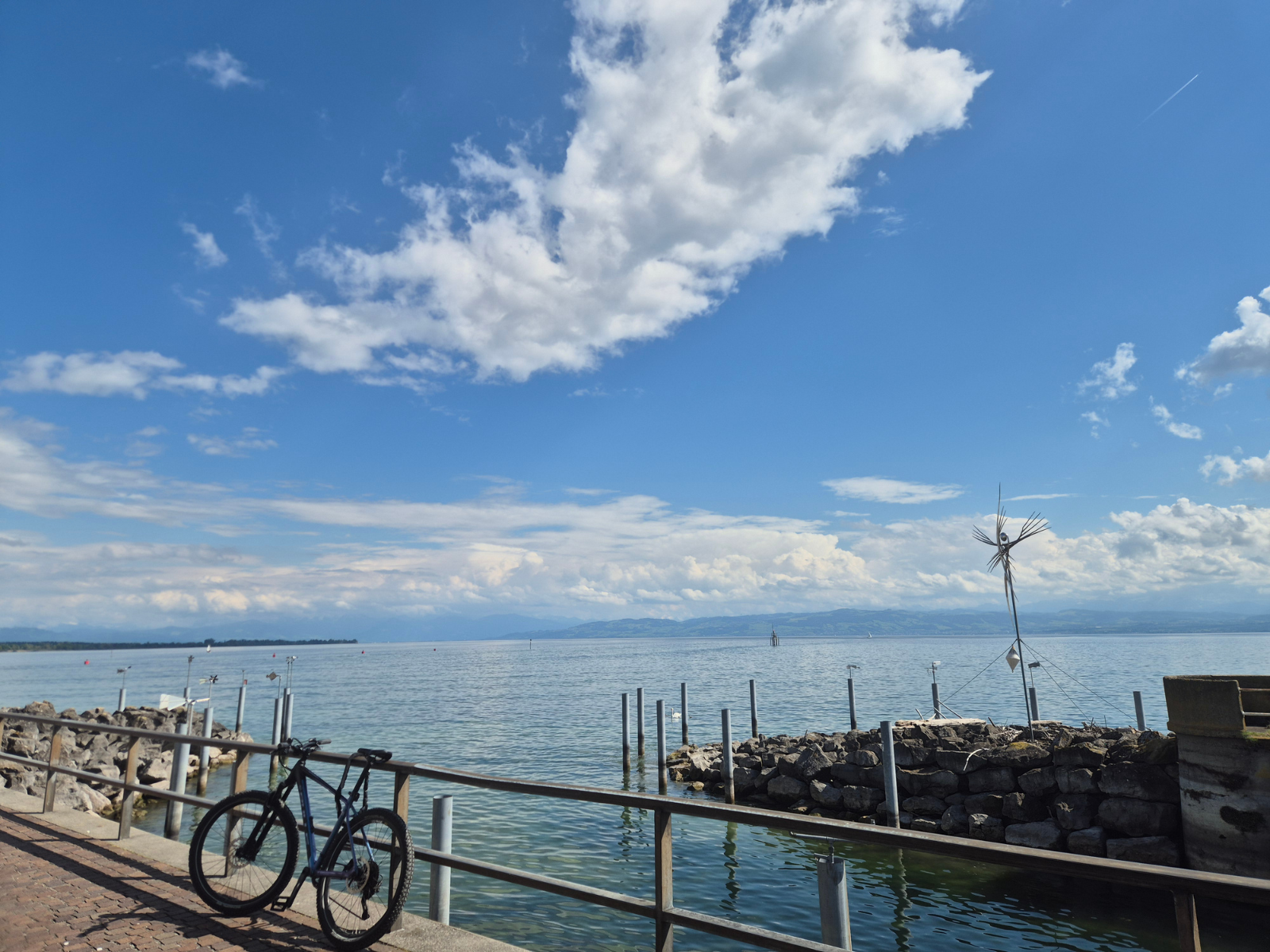 A lone bicycle leaning against a railing on the Friedrichshafen waterfront, Lake Constance calm and wide under a big summer sky.