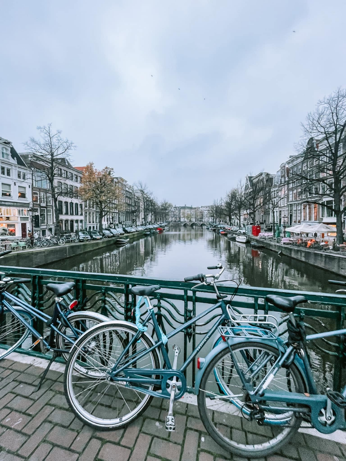 Dutch bicycles locked to a canal bridge railing in Amsterdam, the waterway stretching into the distance between two rows of gabled townhouses on a grey winter day.