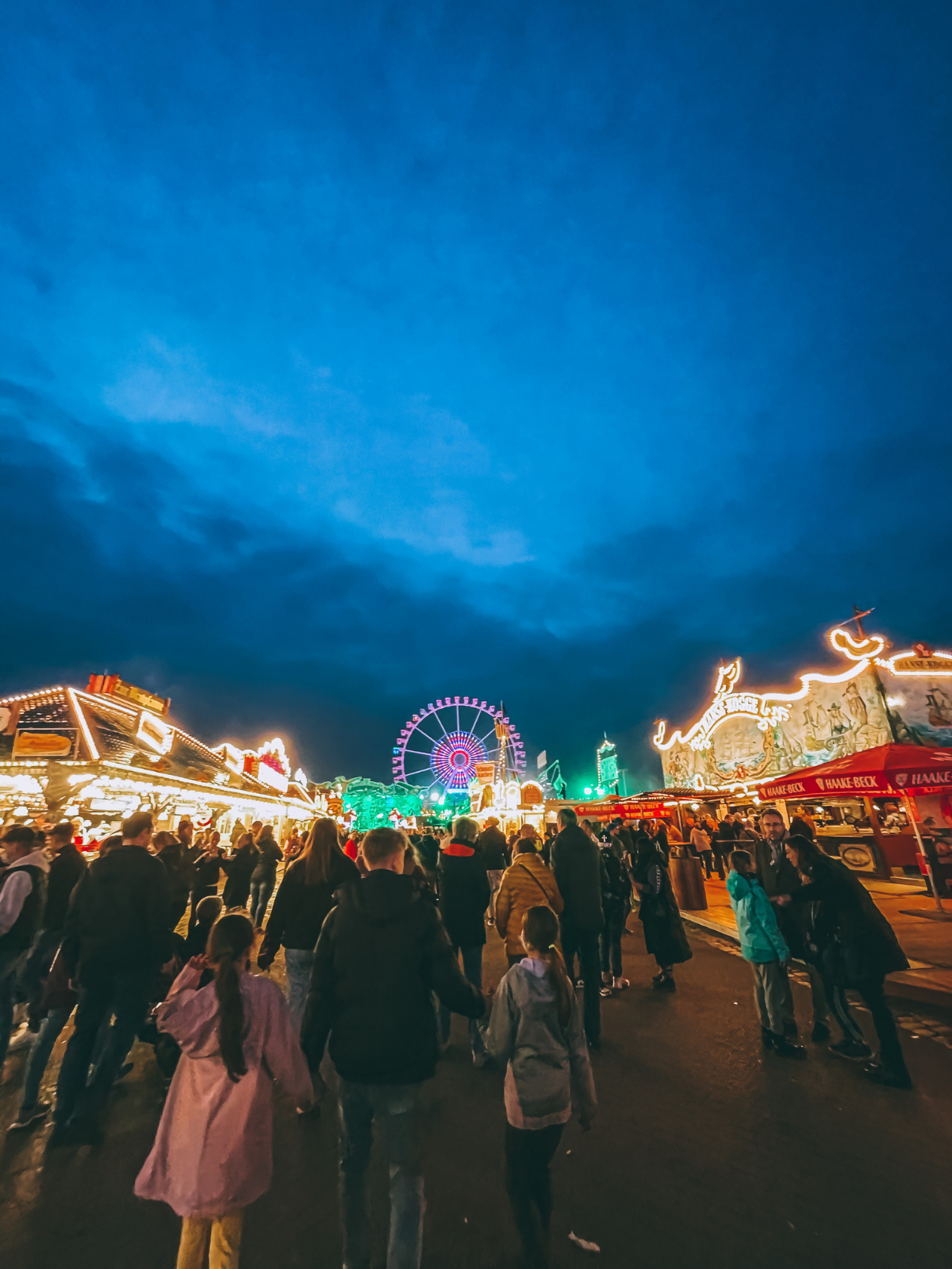 Crowds moving through Bremen's Freimarkt fairground at night, a glowing Ferris wheel and neon fair lights burning against a deep blue sky.
