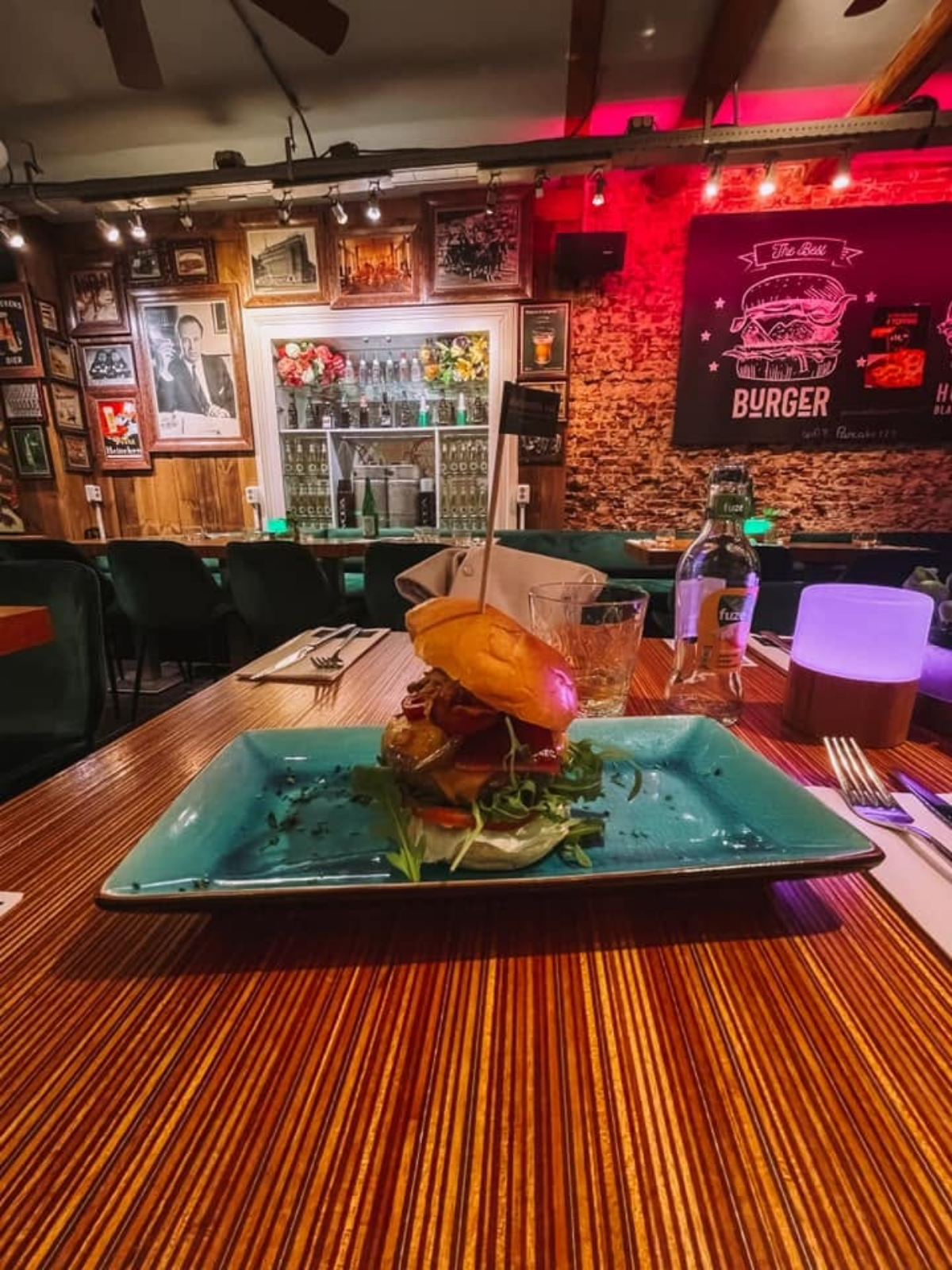 A stacked burger on a turquoise plate at a dimly lit Amsterdam restaurant, framed portraits and exposed brick walls glowing under red neon light.