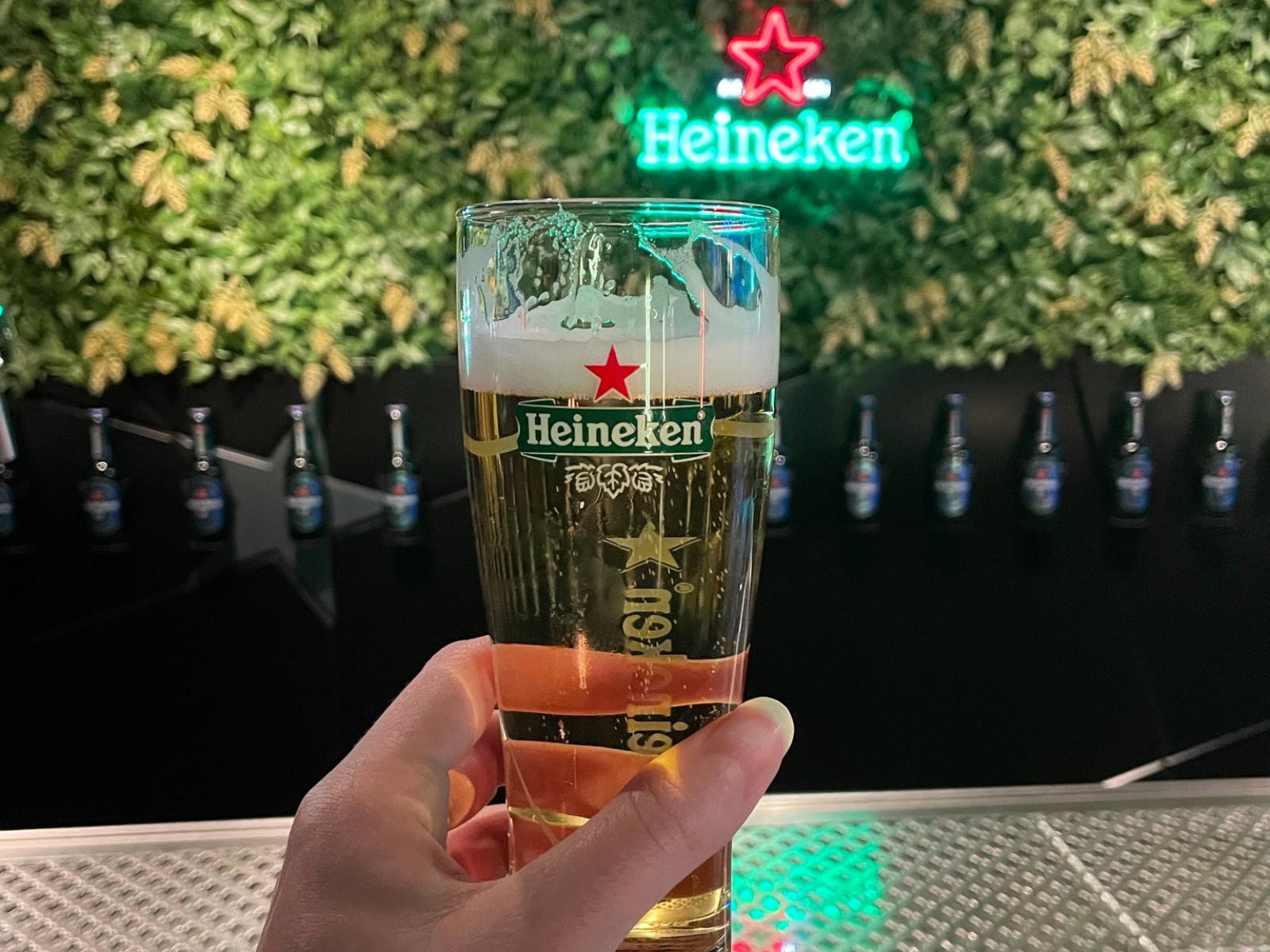 A cold Heineken glass held up in front of the brand's neon sign and green wall installation at the Heineken Experience in Amsterdam.