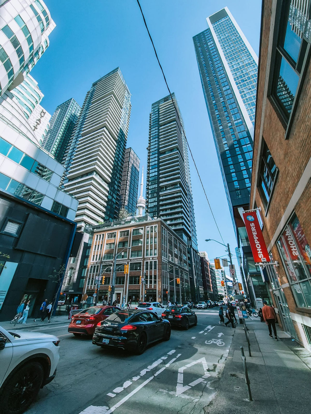 A downtown Toronto street scene with modern high-rise towers, traffic, and the CN Tower visible in the background under a clear blue sky.