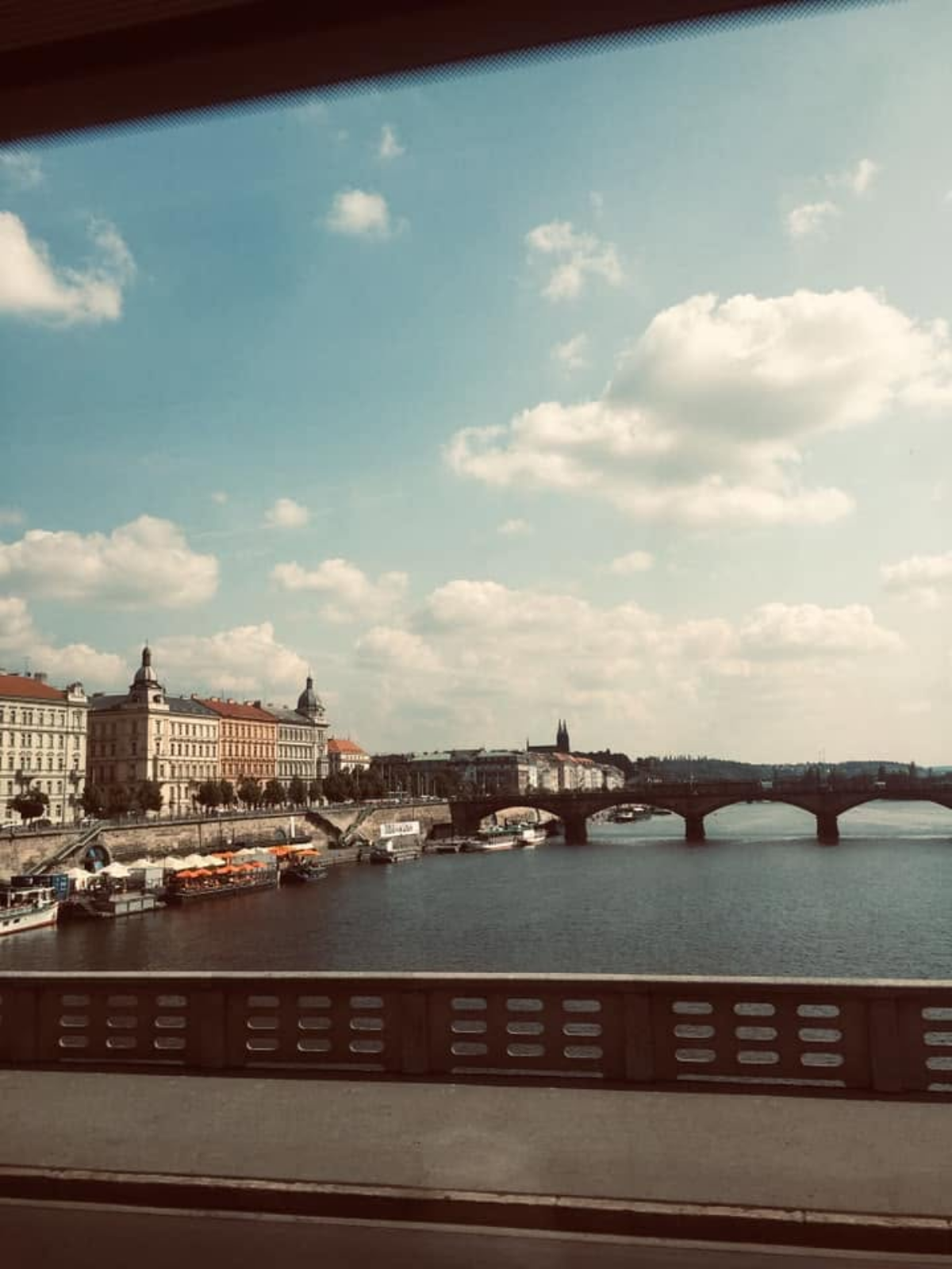 A view of the Vltava River in Prague from a bridge, with riverside buildings, a stone arch bridge, and Prague Castle visible in the distance.