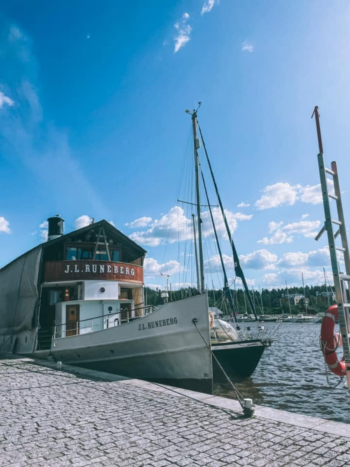 The historic steamship J.L. Runeberg moored at a stone dock in Porvoo, Finland, with sailboats and a marina in the background under a blue sky.