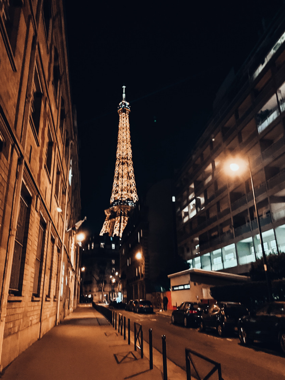 The Eiffel Tower glowing gold at the end of a quiet Parisian side street at night, framed by apartment buildings on either side.