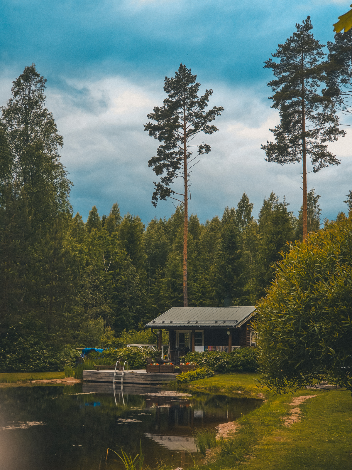 A classic Finnish lakeside cabin nestled between towering pine trees, its dock reflected in the still water on a moody summer evening.