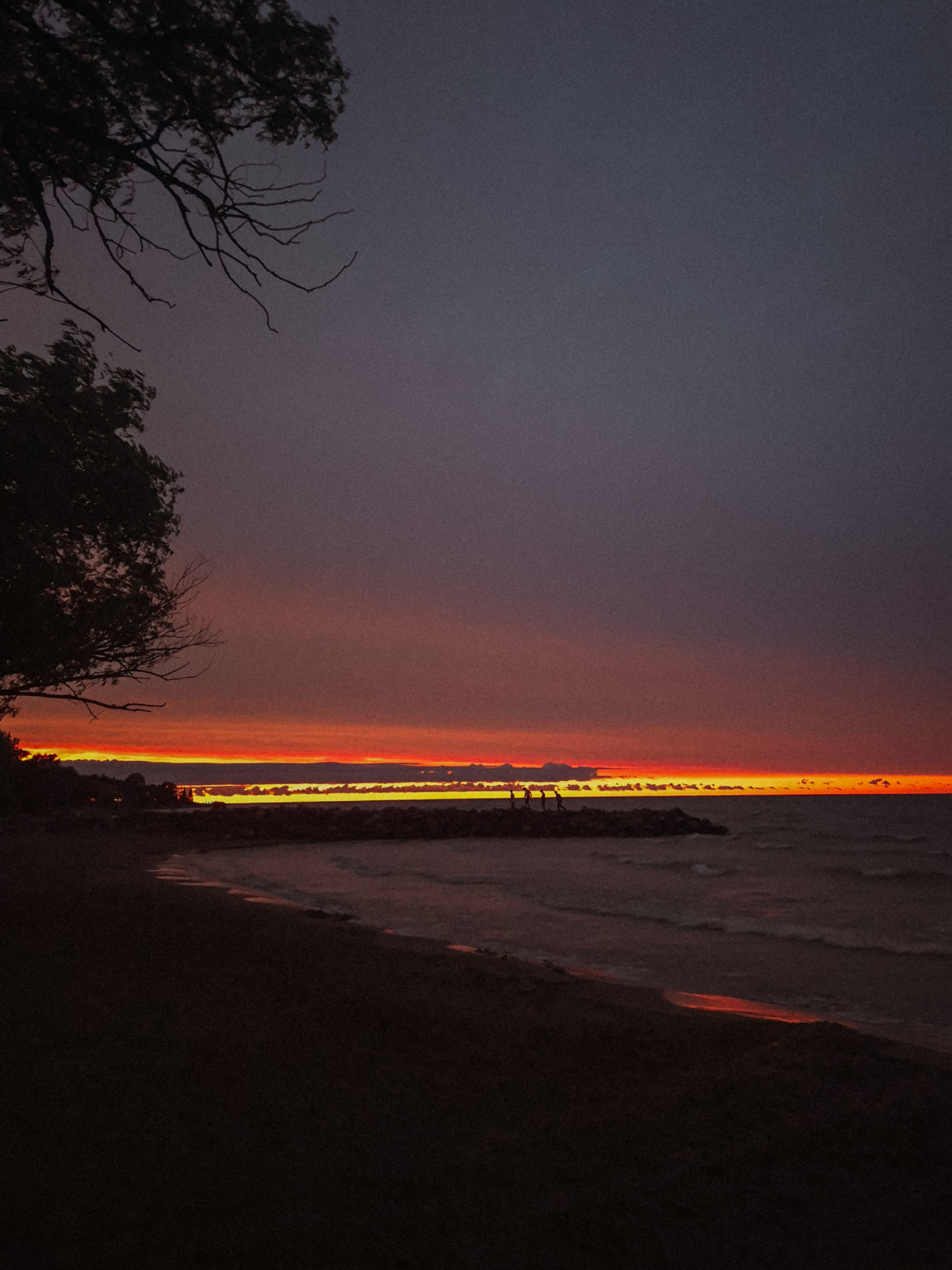 A fiery red and orange band of sunset burning low over Georgian Bay, the dark shoreline and silhouetted trees framing the last light of the day.