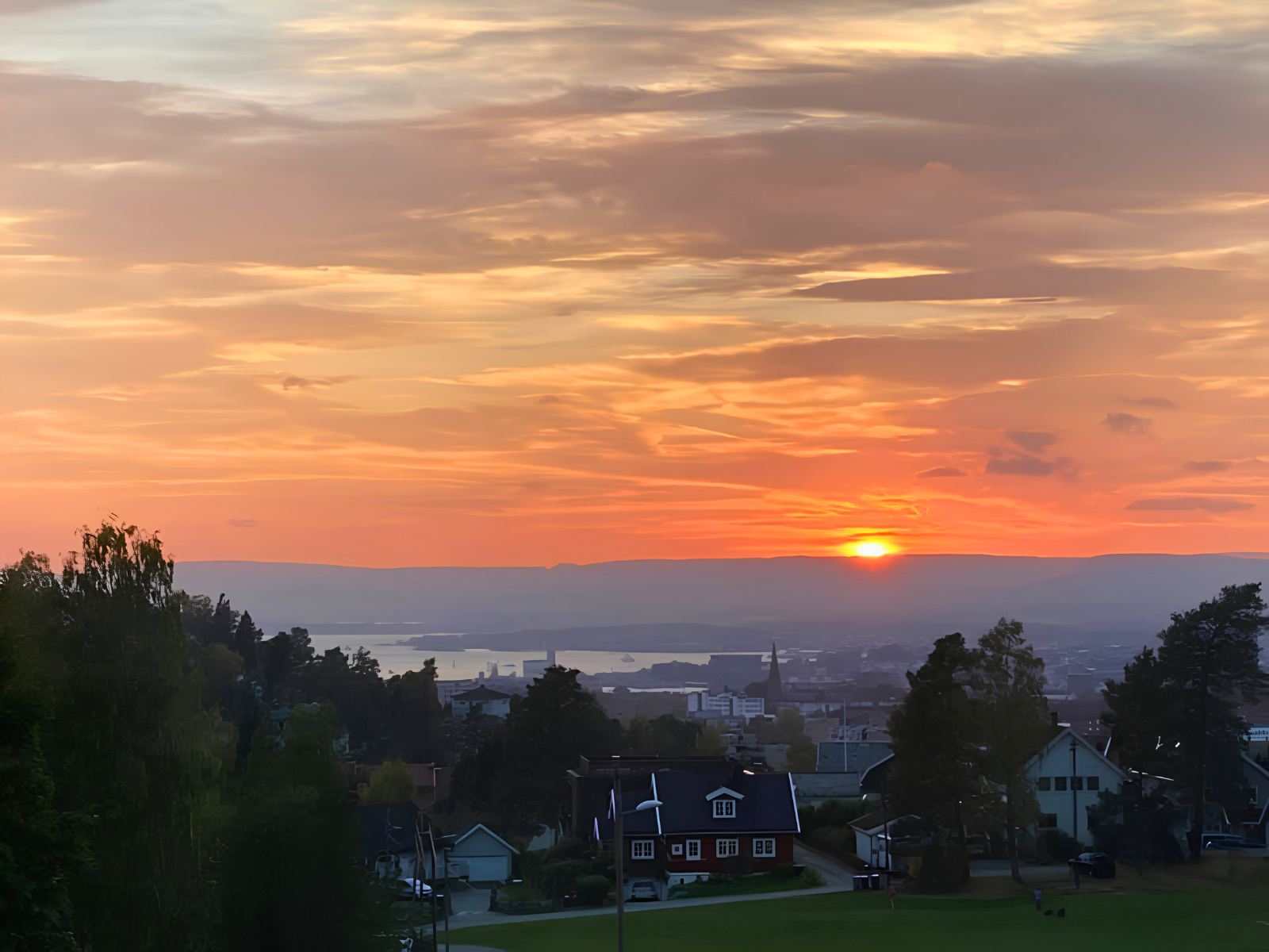 A blazing orange and pink sunset over the Oslo fjord seen from the hillside, the city skyline and church spire silhouetted below.