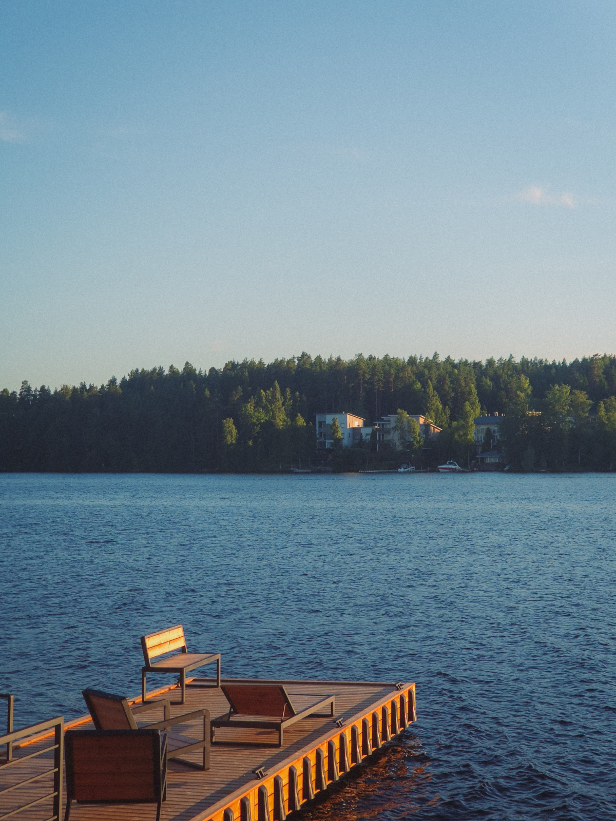 Two wooden chairs on a glowing lakeside dock at golden hour in Finland, the still water and pine forest stretching out across the horizon.