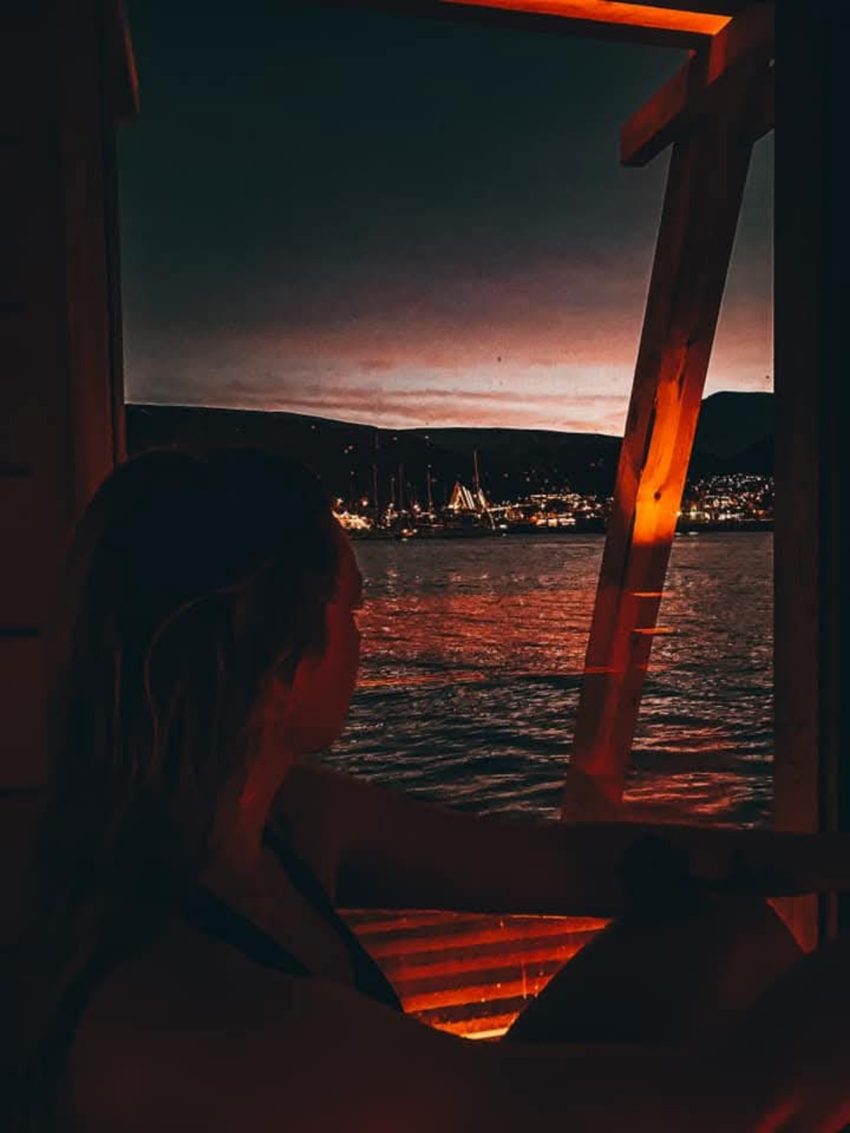 A silhouette looking out through a sauna window toward the glowing lights of Tromsø across the water at dusk, warm amber light against the dark.