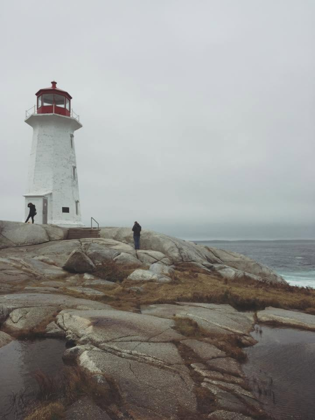 The white and red Peggy's Point Lighthouse on the rocky coastline of Peggy's Cove, Nova Scotia, under an overcast sky with the ocean in the distance.
