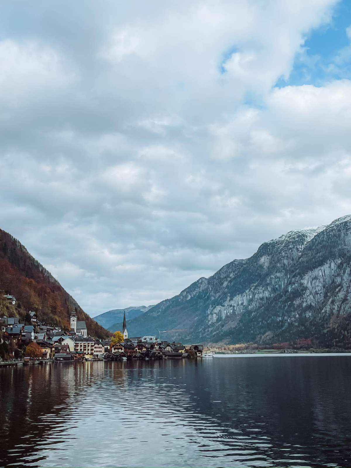 Hallstatt village reflected in the still dark waters of the Hallstättersee, its church spire and alpine rooftops dwarfed by the sheer grey cliff face rising behind.