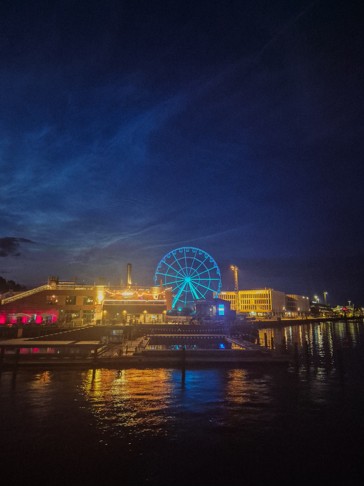 A glowing blue Ferris wheel reflected in the dark water of Helsinki's harbour at night, the waterfront lit in gold and red.