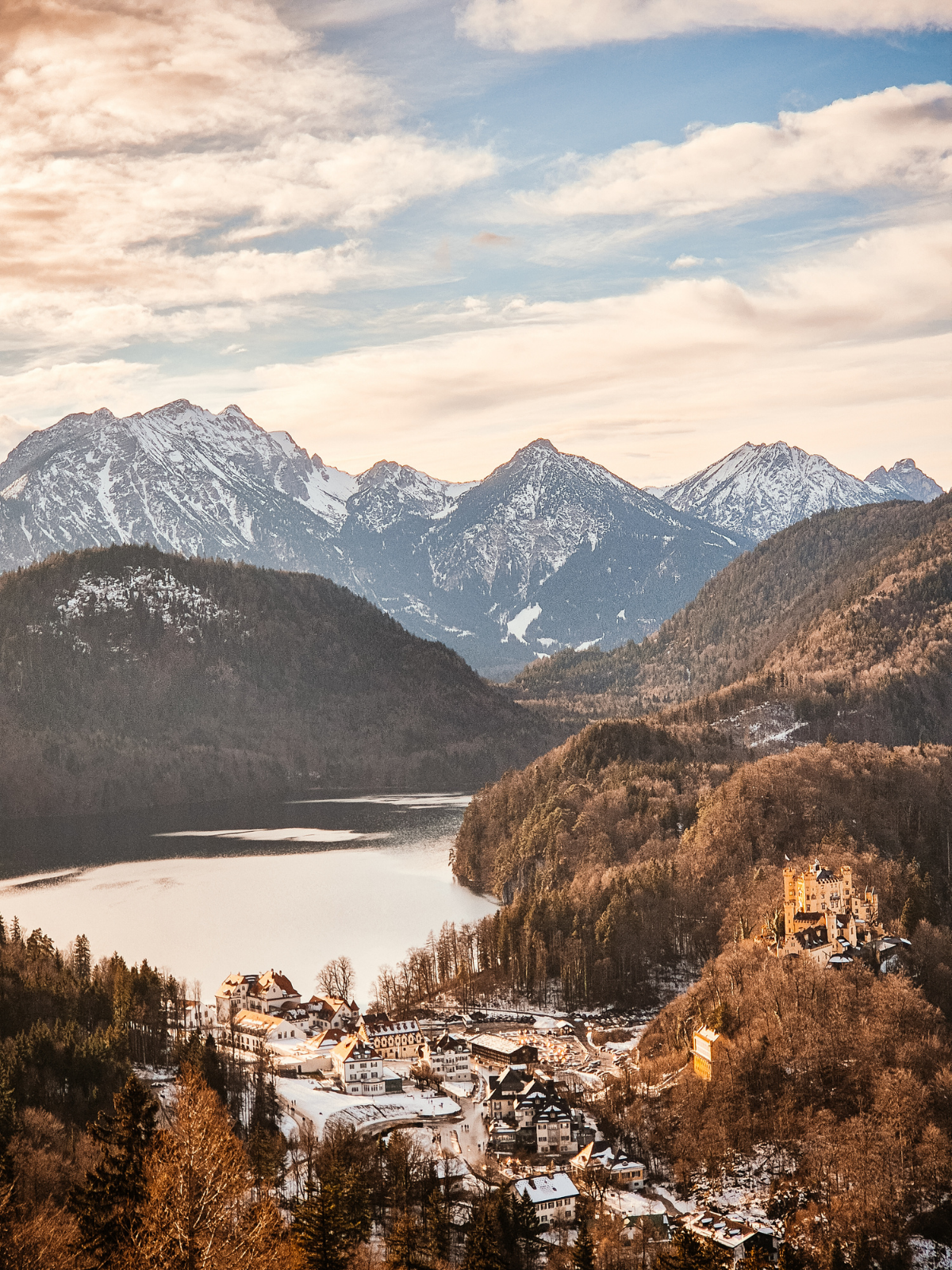 A golden winter panorama from Neuschwanstein looking down over Hohenschwangau village, the frozen Alpsee lake, and snow-dusted Bavarian Alps at dusk.