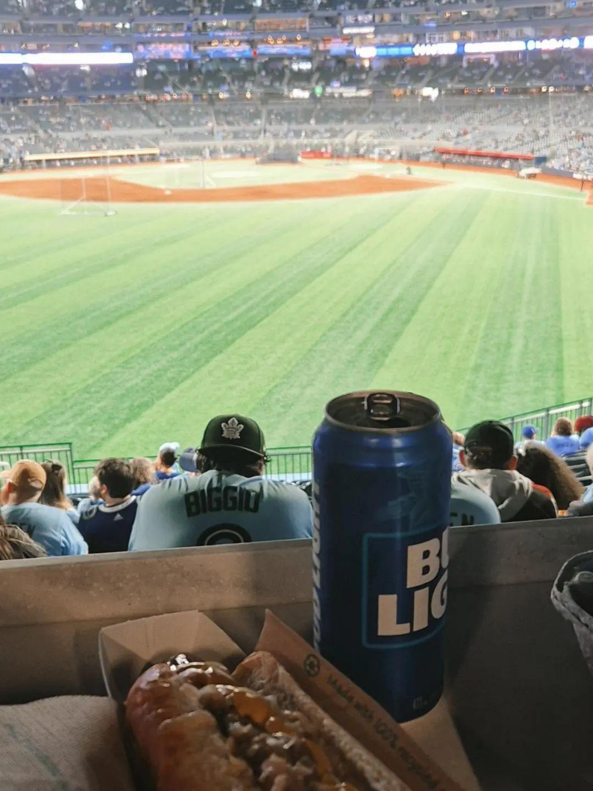 A Bud Light can and a loaded hot dog in the foreground, the glowing green diamond of a Toronto Blue Jays game stretching out behind.