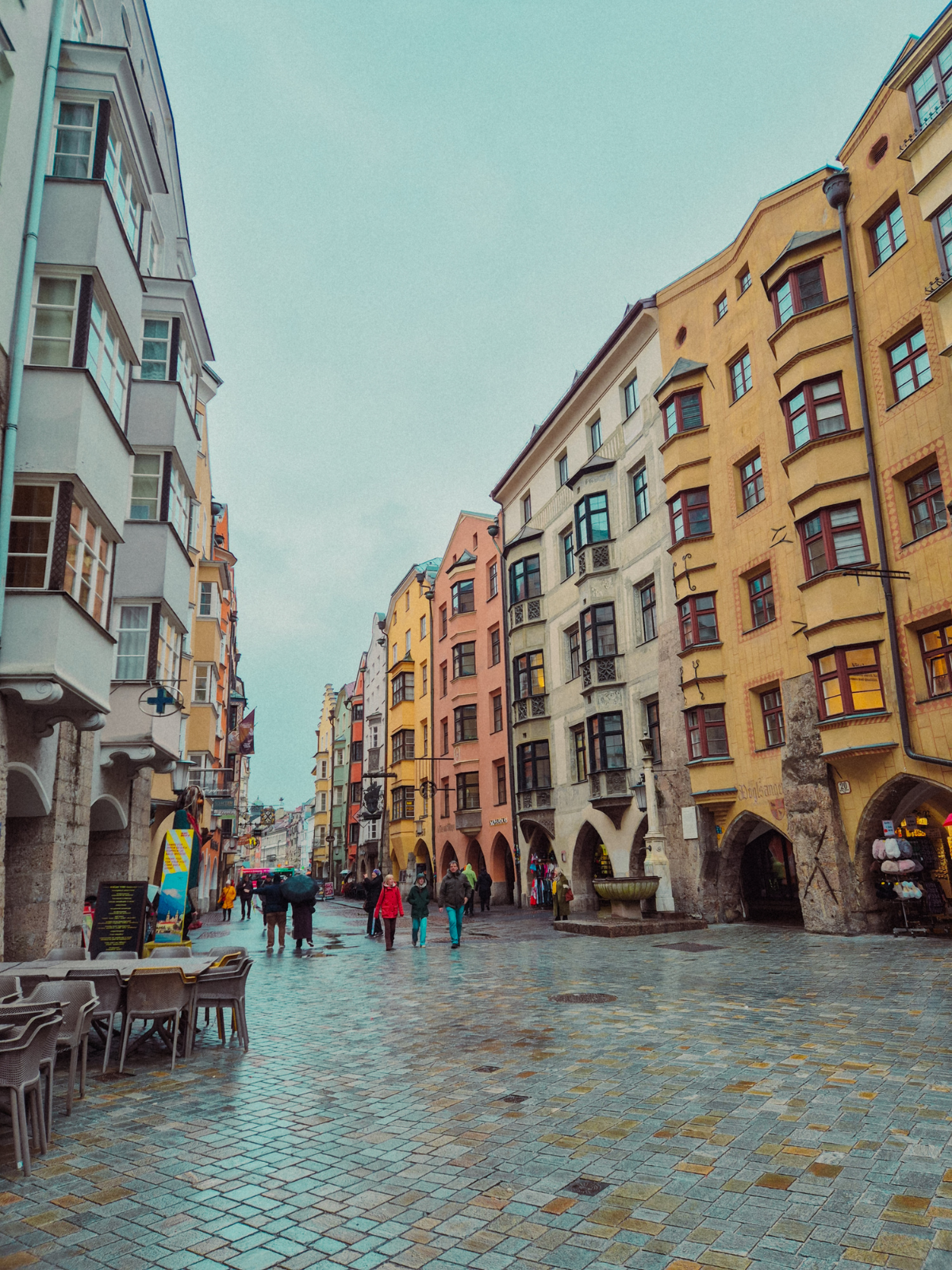 The colourful medieval facades of Innsbruck's Herzog-Friedrich-Strasse on a rainy afternoon, warm window light glowing behind arched arcades.