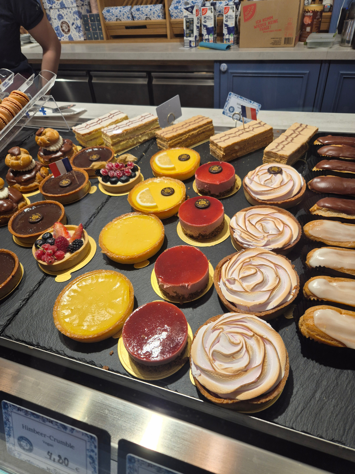 A French patisserie display case in Ravensburg, lined with lemon tarts, raspberry mousse cakes, meringue roses, and chocolate éclairs on slate trays.