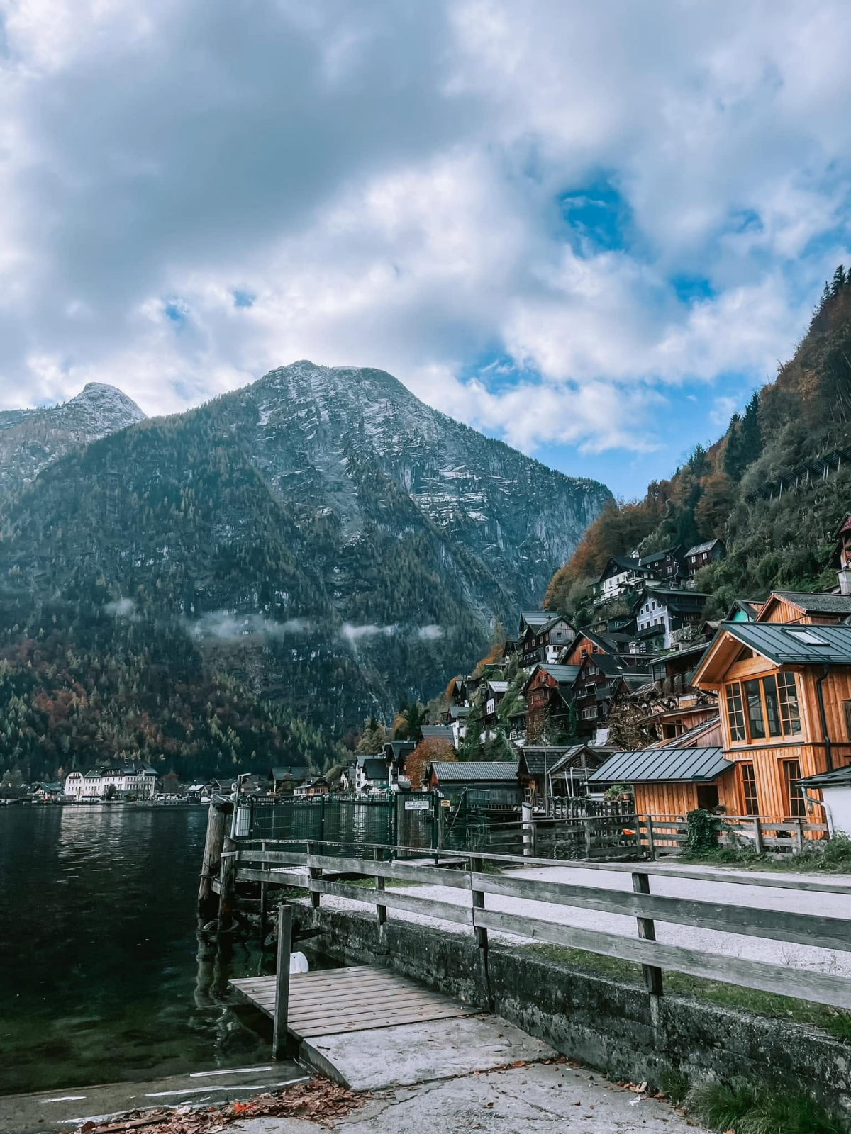 Hallstatt's timber lakeside chalets and boat houses clustered at the foot of a towering rock face, viewed from a wooden dock along the water's edge in autumn.