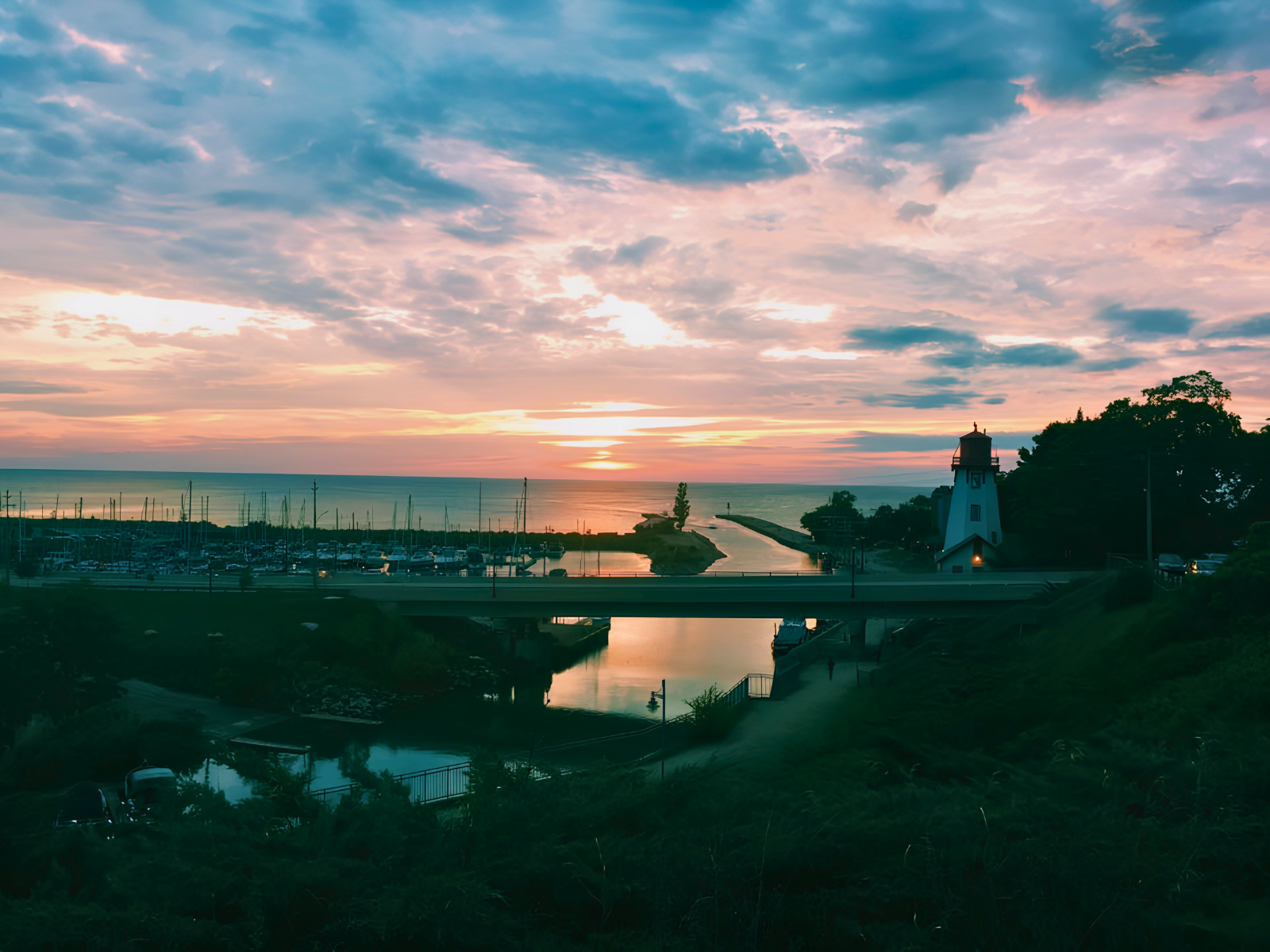 A pastel sunset over an Ontario marina, a white lighthouse glowing at the harbour entrance, sailboat masts silhouetted against the fading sky.