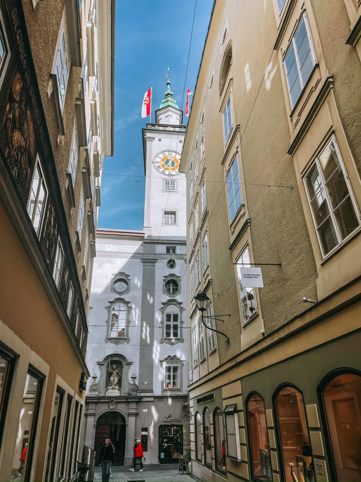 A narrow historic alley in Salzburg, Austria, framed by pastel buildings and leading up to a baroque clock tower with Austrian flags.