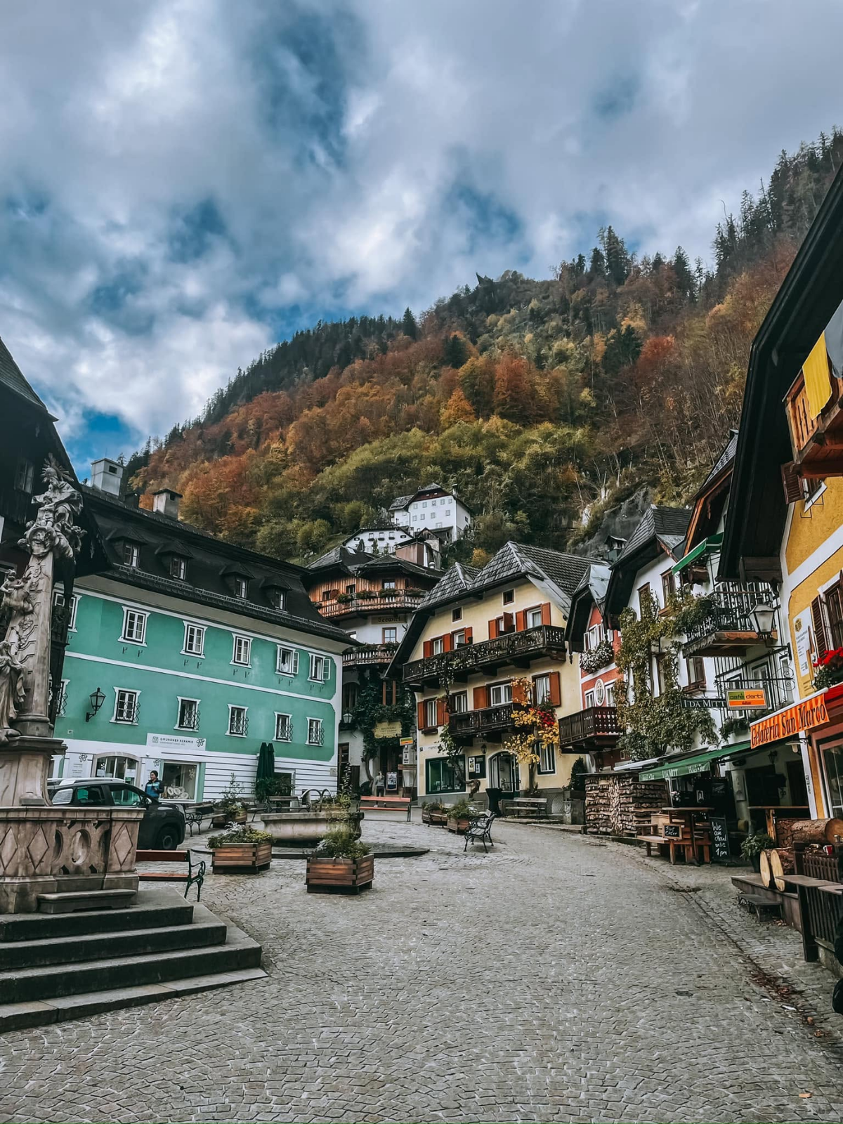 Hallstatt's cobblestone market square framed by colourful alpine buildings and a hillside ablaze with autumn colour rising sharply behind.