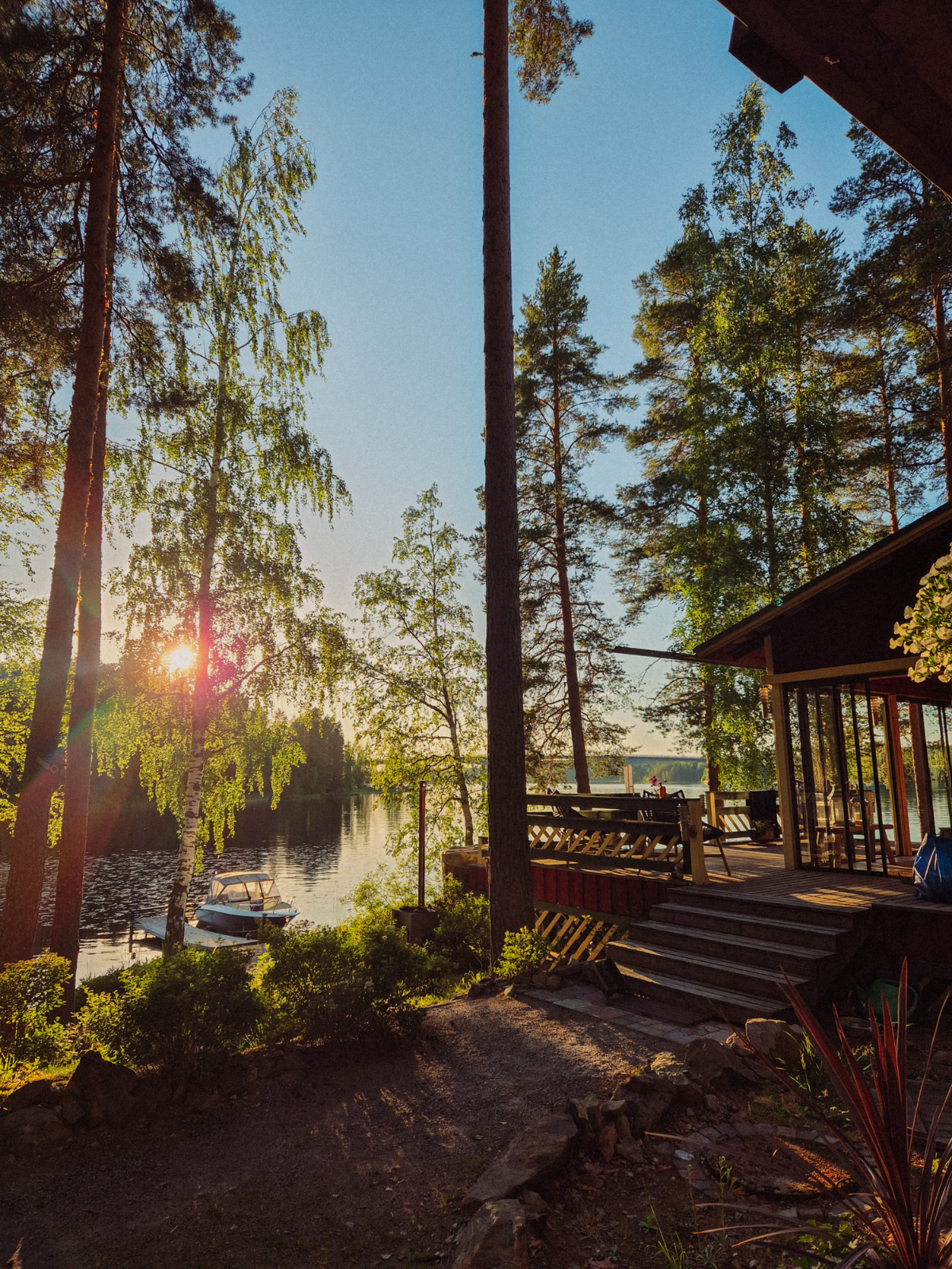 The sun setting through birch trees at a Finnish lakeside cabin in Kalkkinen, a small boat moored at the dock in the warm midsummer light.