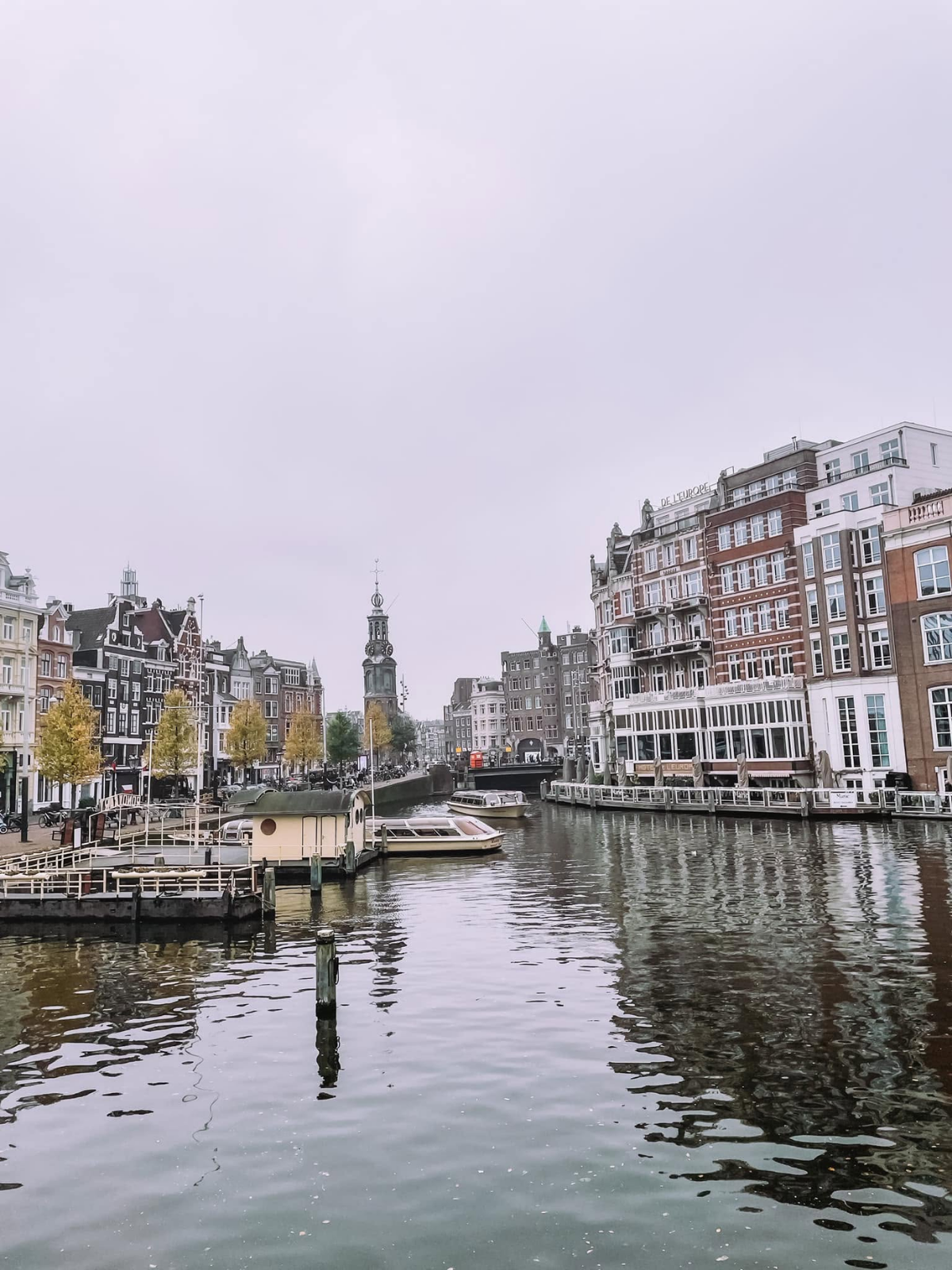 A wide Amsterdam canal on an overcast autumn day, the Munttoren clock tower rising in the distance between historic Dutch townhouses and a moored houseboat.