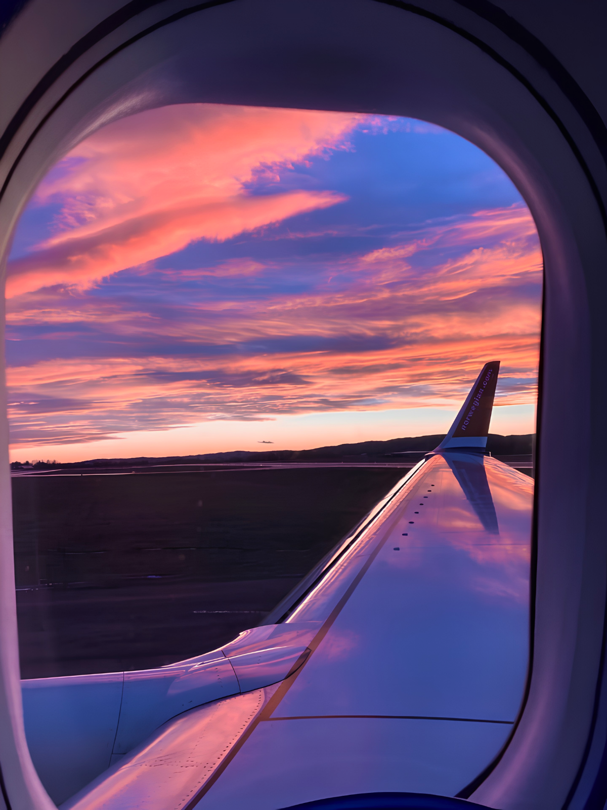 A Norwegian Air wing cutting through a dramatic pink and violet sunset sky, viewed through an oval airplane window on the tarmac.