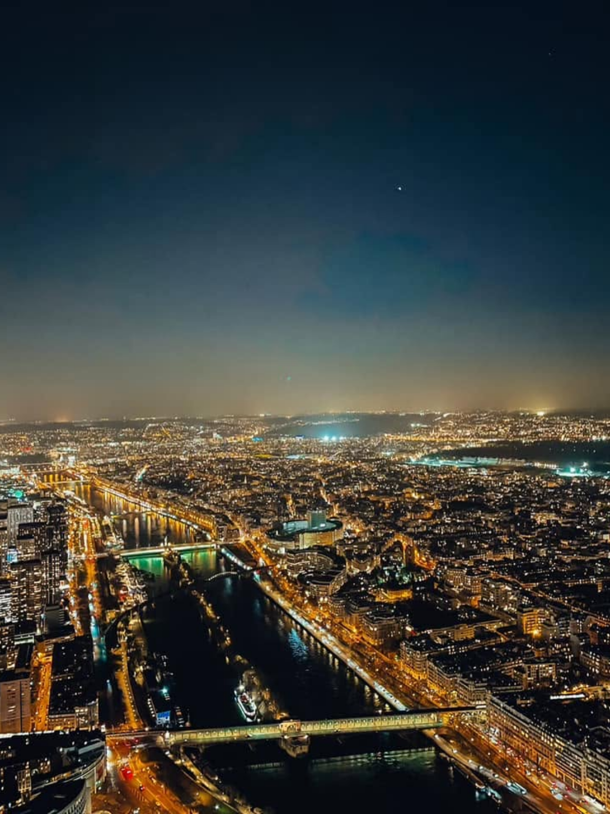 A night view of Paris from an elevated vantage point, showing the illuminated Seine River winding through the city with bridges and lit streets below.