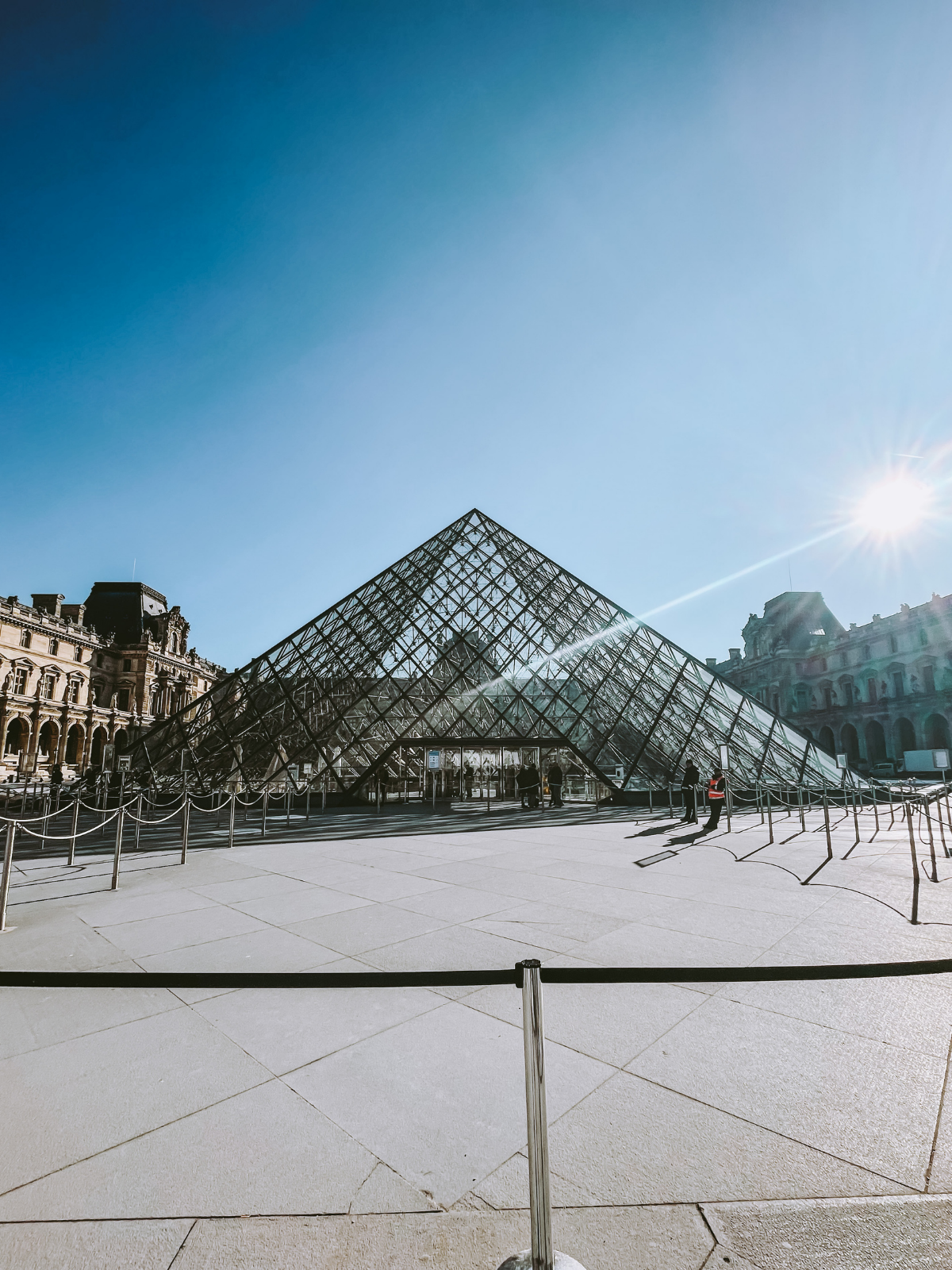 The Louvre glass pyramid catching the morning sun against a brilliant blue Paris sky, the courtyard nearly empty and sharp with winter light.