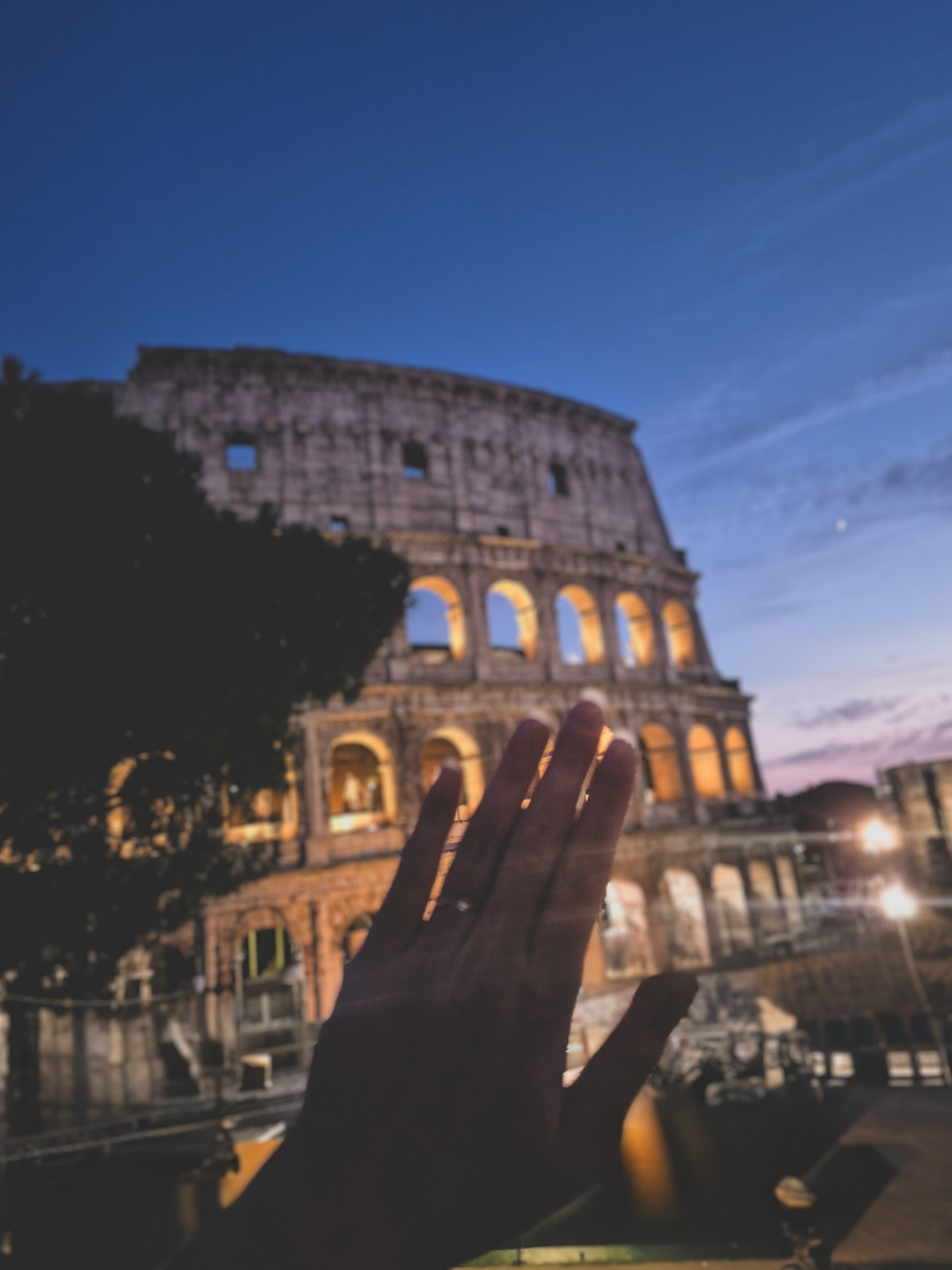 A hand bearing a new engagement ring reaches toward the illuminated Colosseum at dusk.