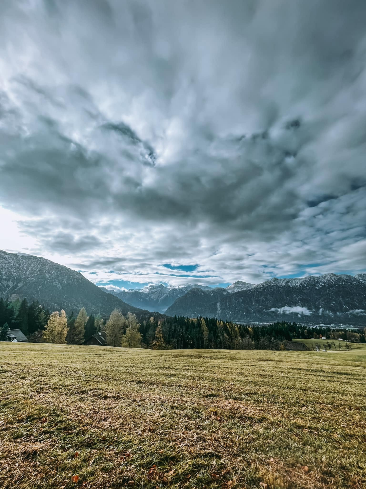 A dramatic sweep of storm clouds rolling over a wide Austrian alpine meadow, snow-capped peaks visible in the blue distance beyond the treeline.