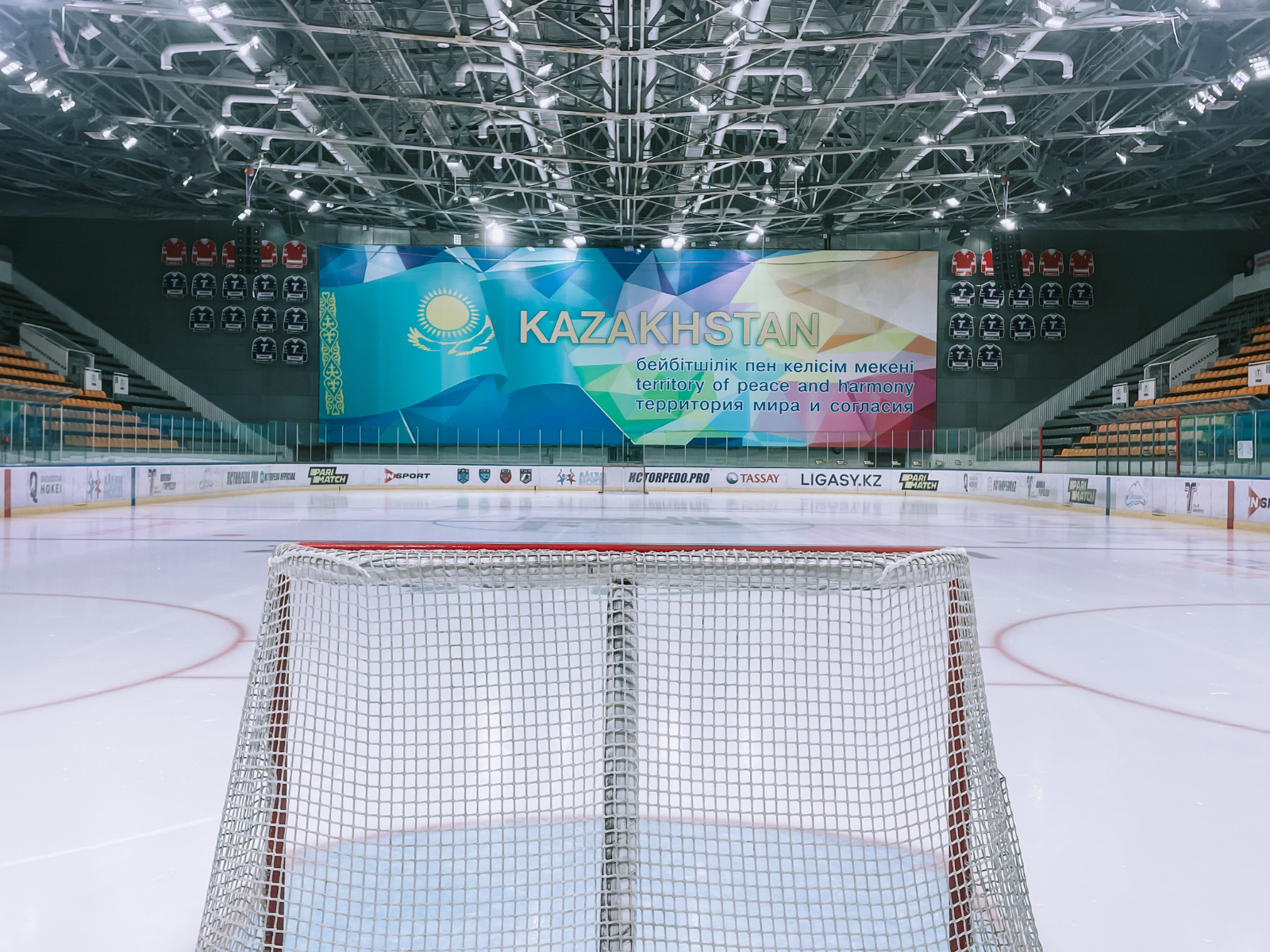 Looking down the ice from behind a hockey net in an empty Kazakhstani arena, the Kazakhstan national banner lit up on the scoreboard.