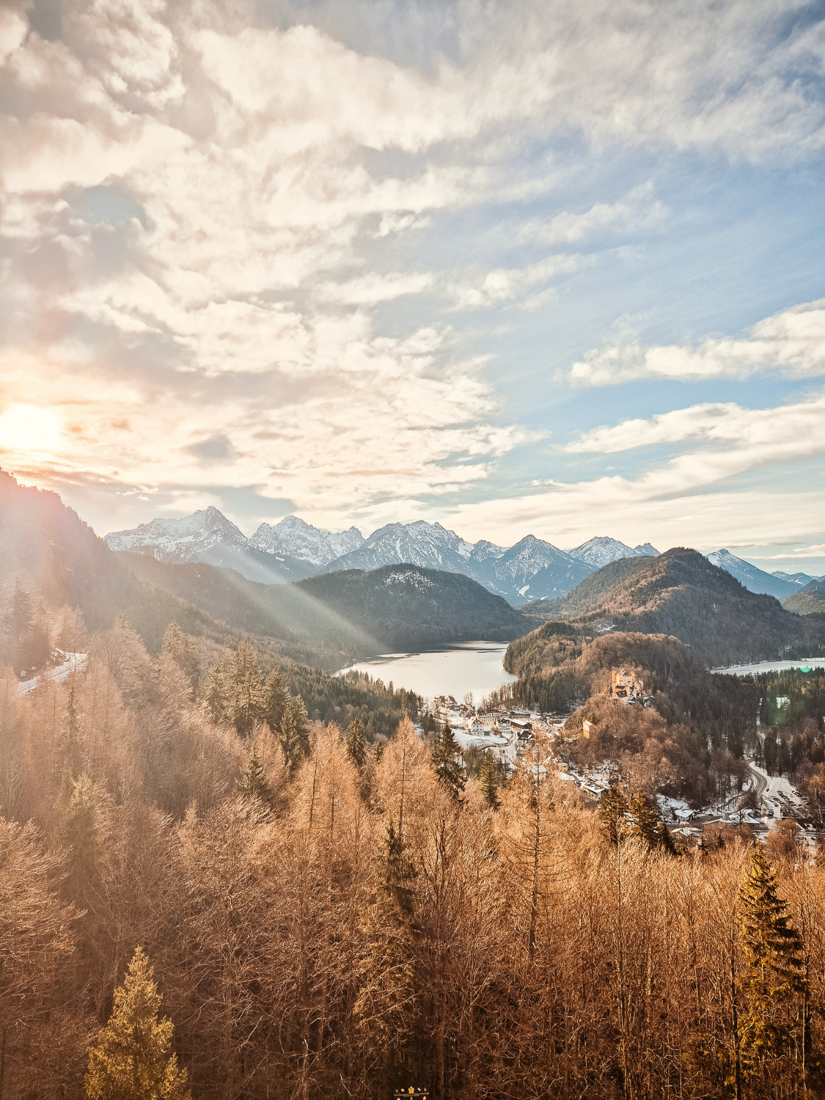 A sweeping Bavarian valley view from Neuschwanstein, golden light breaking through winter clouds over snow-capped Alps and a frozen lake below.