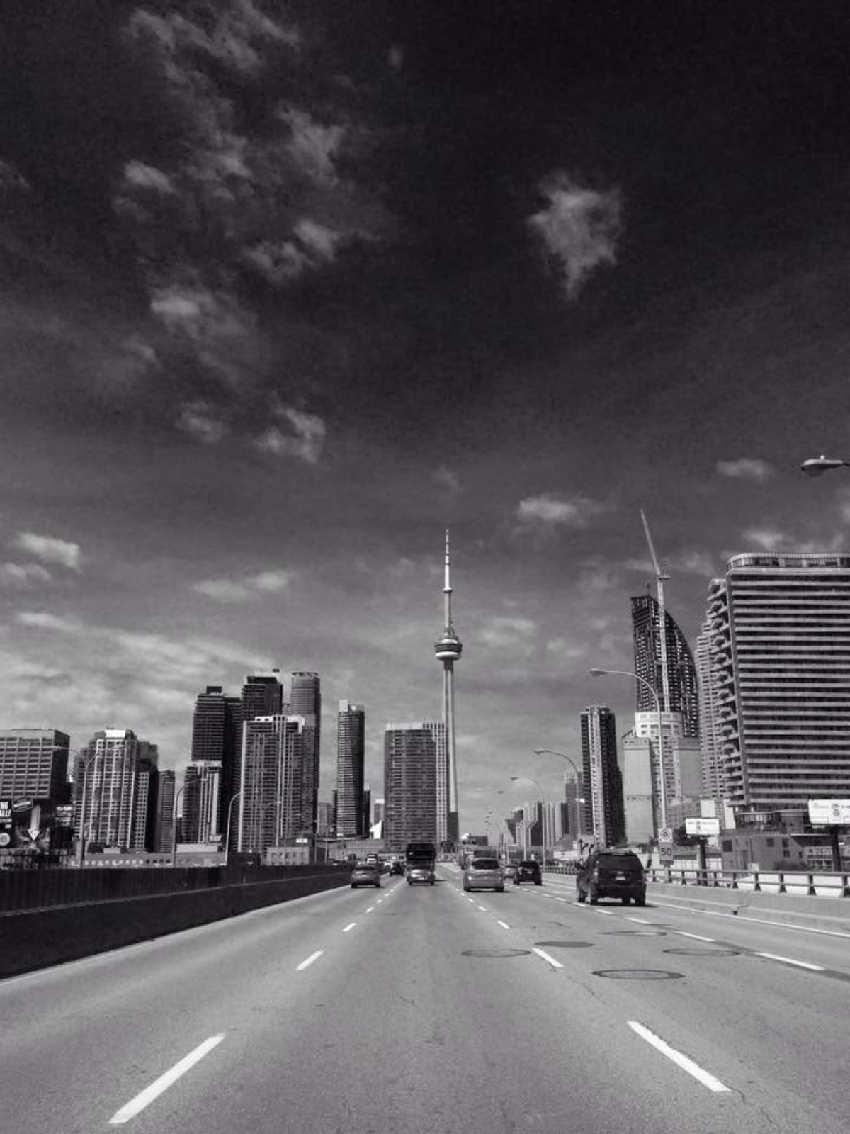 A black-and-white photo of the Toronto skyline and CN Tower, shot from a highway with cars driving toward the city under a dramatic cloudy sky.