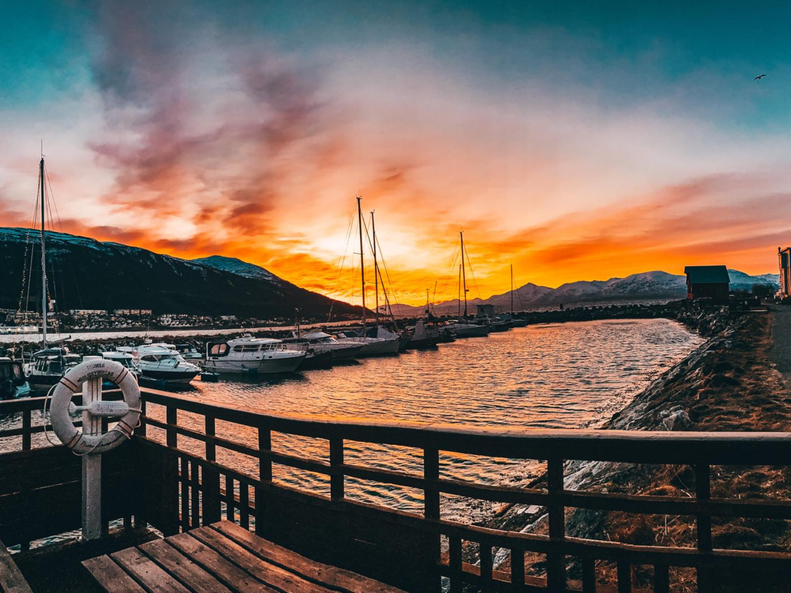 A blazing orange and teal sunset over Tromsø harbour, sailboat masts silhouetted against the burning sky, a life ring on the dock railing in the foreground.