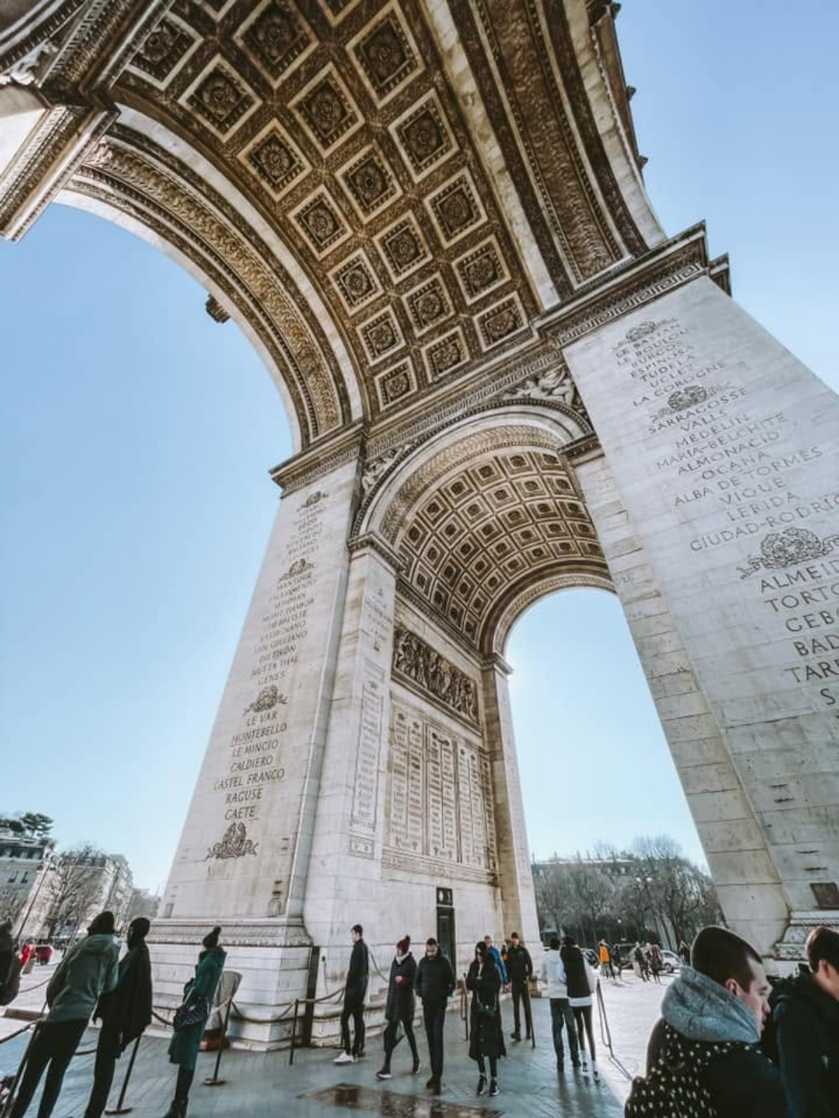 Looking up from directly beneath the Arc de Triomphe in Paris, its ornate coffered ceiling and carved stone pillars towering overhead against a clear blue sky.