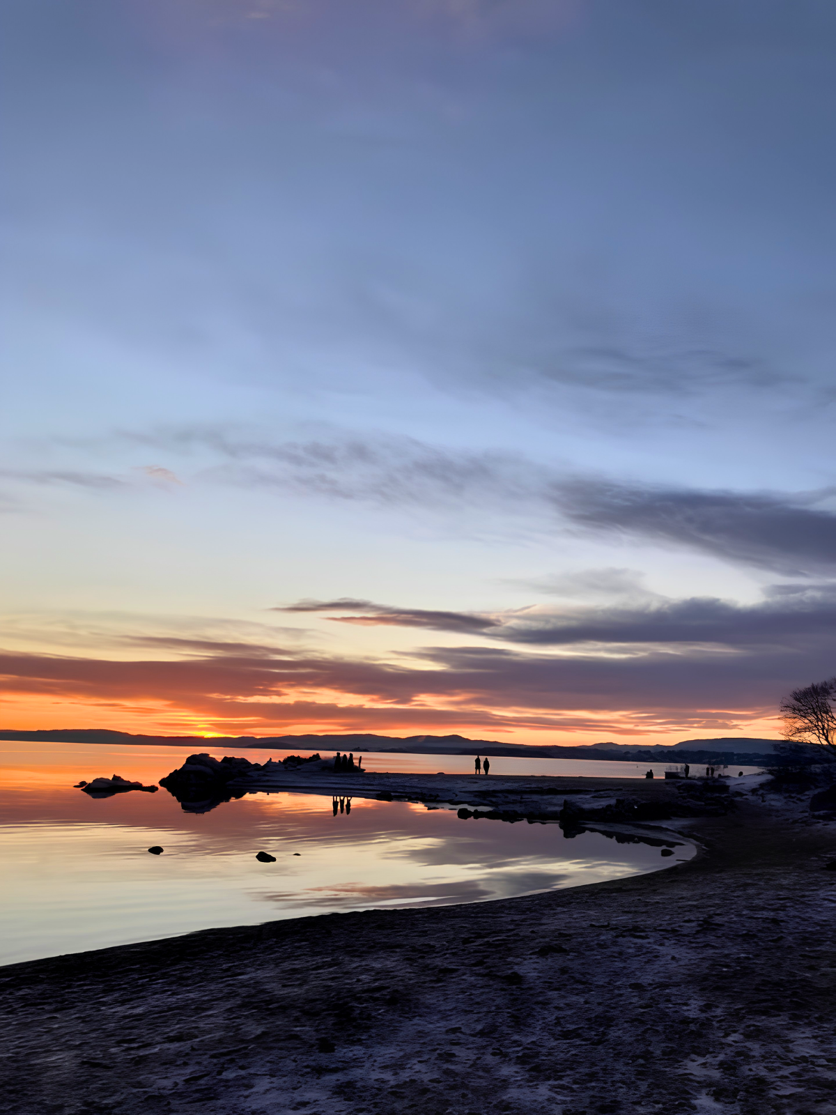 A still winter sunset over the Oslofjord, the orange horizon reflected perfectly in glassy water, silhouetted figures standing at the shoreline.